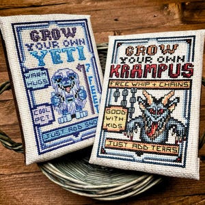 Pode incluir: Duas amostras de ponto cruz numa bandeja tecida. Uma diz "Grow Your Own Yeti" com um yeti de desenho animado. A outra diz "Grow Your Own Krampus" com uma ilustração de Krampus. Ambas têm texto e são emolduradas em azul.