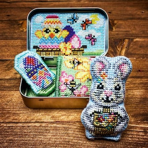 May include: A set of four small cross-stitch embroidery pieces in a tin box. The pieces feature a bunny, a chick, flowers, and a bee. The tin box is decorated with a floral design.