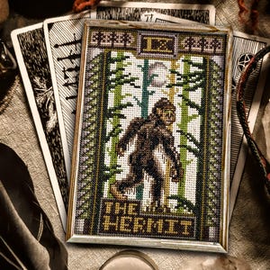 May include: A framed cross-stitch tarot card showing "The Hermit" with a Bigfoot-like figure in a forest setting. The card is displayed with other tarot cards and mystical items, using green, brown, and beige tones.