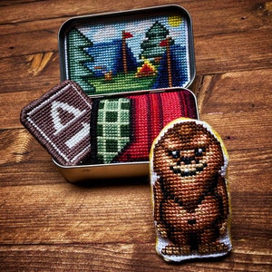 May include: A metal tin with cross-stitched patches. The patches include a camping scene, a brown patch with the letter "A", a green and black plaid patch, a red striped patch, and a brown Bigfoot patch.