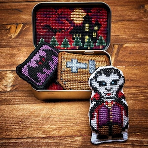 Può includere: Un set di quattro ornamenti a punto croce a tema Halloween. Gli ornamenti sono in una piccola scatola di latta. Gli ornamenti includono un pipistrello viola, una bara marrone con una croce bianca e un vampiro bianco con un mantello rosso e una camicia viola.