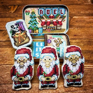 Könnte beinhalten: Eine Sammlung handgefertigter Weihnachtsartikel aus Kreuzstich. Enthält drei Weihnachtsmannfiguren, eine Dose mit dem gestickten Schriftzug "NOEL" und einen Panda mit Geschenken. Die Artikel befinden sich auf einer Holzoberfläche.