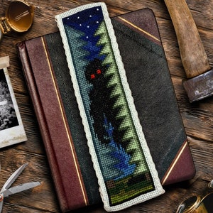 May include: A cross-stitch bookmark with a dark blue background and a black silhouette of a creature with red eyes standing in a forest. The bookmark is surrounded by a white lace border.