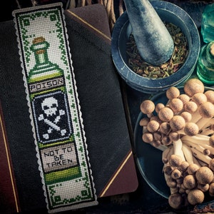 Peut inclure: Marque-page en point de croix avec une bordure verte et blanche, représentant un flacon de poison avec le mot "POISON" et une tête de mort et des os croisés. Le texte "NOT TO BE TAKEN" est également visible. L'image comprend un livre, des champignons et des articles d'apothicaire.