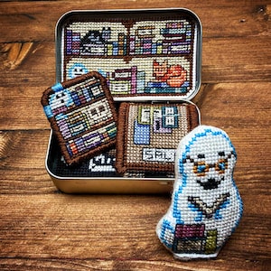 Może przedstawiać: Zestaw czterech małych, kolorowych, haftowanych filcowych ozdób w kształcie książek w blaszanym pudełku. Ozdoby mają kształt książek i są w różnych kolorach i wzorach. Jedna ozdoba ma kształt ducha w okularach i trzyma książkę.