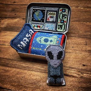 Può includere: Una scatola di latta ricamata blu e bianca con un'astronave e la parola "NASA" su di essa. La scatola è aperta e un giocattolo di peluche alieno grigio è seduto di fronte ad essa.