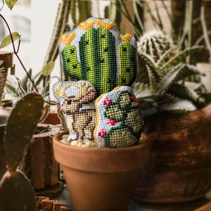 Könnte beinhalten: Ein Set aus drei bestickten Kaktus-Kissen in einem Terrakotta-Topf. Die Kissen sind grün, gelb und braun mit einem Hirsch-Design.