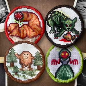 Peut inclure: Quatre patchs brodés représentant des créatures mythologiques. Les patchs sont circulaires et ont un fond blanc avec des broderies colorées. Le premier patch représente une créature brune ressemblant à Bigfoot debout dans une forêt. Le deuxième patch représente un monstre vert avec des yeux rouges et des griffes. Le troisième patch représente un ours brun avec une fleur rouge dans sa patte. Le quatrième patch représente une créature verte avec des yeux rouges et une bouche rouge.