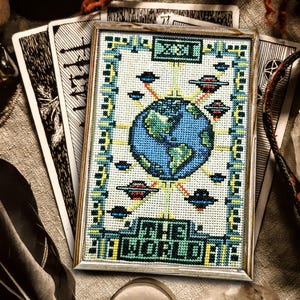 May include: A framed cross-stitch showing the Earth with UFOs and the text "THE WORLD". The design uses blue, green, yellow, and red. Tarot cards and other mystical items are in the background, creating a mystical theme.
