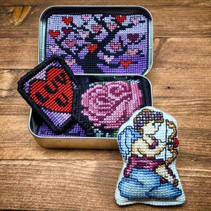 Könnte beinhalten: Eine Metallbox mit Kreuzstichkunst, darunter ein Baum mit herzförmigen Blättern, ein herzförmiger Aufnäher mit "LUV U", eine rosa Rose und eine Amorfigur. Die Artikel liegen auf einer Holzoberfläche.