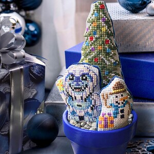 May include: A blue pot with a green, white, and blue Christmas tree, a white snowman, and a blue and white Yeti ornament. The pot is sitting on a table with a blue and silver gift box in the background.