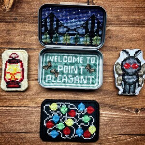 Op de afbeelding: Vier geborduurde patches met een lantaarn, een mot, een lichtsnoer en een blik met de tekst "Welkom in Point Pleasant".