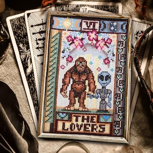 Könnte beinhalten: Kreuzstich-Kunstwerk der Tarotkarte "Die Liebenden", mit einer Bigfoot-ähnlichen Figur und einem Alien. Das gerahmte Stück hat einen hellen Holzrahmen und ist von Tarotkarten und Kristallen umgeben. Das Design enthält florale und himmlische Elemente.