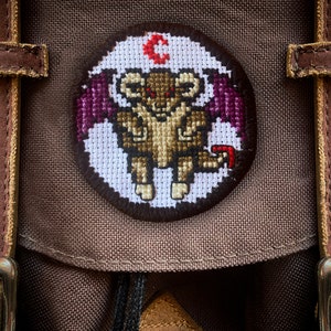 May include: A brown canvas backpack with a circular embroidered patch featuring a purple-winged, red-eyed, brown creature with the letter "C" above it.