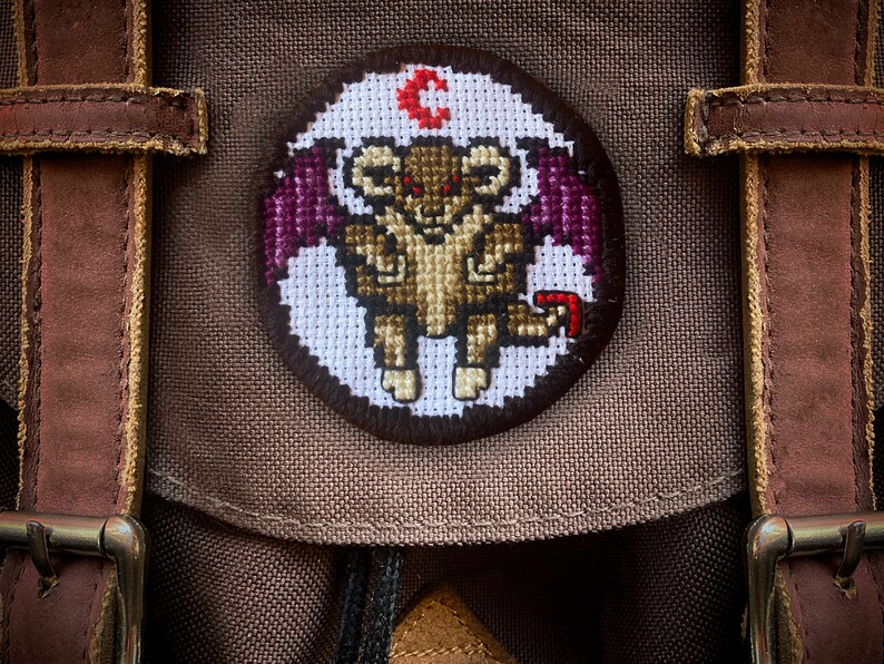 May include: A brown canvas backpack with a circular embroidered patch featuring a purple-winged, red-eyed, brown creature with the letter "C" above it.
