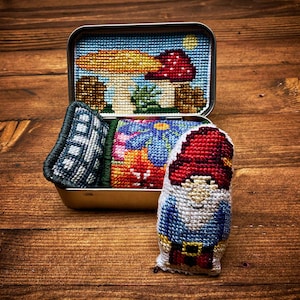 Może przedstawiać: Mały, ręcznie szyty gnom z czerwonym kapeluszem, niebieską koszulą i białą brodą. Gnom siedzi obok blaszanego pudełka z kolorowym wzorem haftu krzyżykowego przedstawiającym grzyb i łóżko.