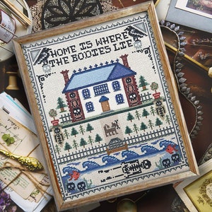 May include: A framed cross-stitch design with a spooky house and the text "Home is where the bodies lie". The house is blue and white with a red brick chimney. There are trees and a fence around the house. The background is a blue and white wave pattern with skeletons and skulls.
