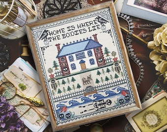 Puede incluir: Un diseño de punto de cruz enmarcado con una casa espeluznante y el texto "Home is where the bodies lie". La casa es azul y blanca con una chimenea de ladrillo rojo. Hay árboles y una valla alrededor de la casa. El fondo es un patrón de olas azul y blanco con esqueletos y calaveras.