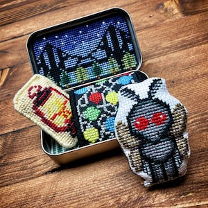 Puede incluir: Un conjunto de cuatro piezas pequeñas bordadas en una lata de metal. Las piezas incluyen una linterna, una escena nocturna, una bombilla y una polilla con ojos rojos.