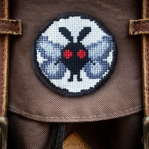 May include: A brown canvas backpack with a circular embroidered patch featuring a black and white moth with red eyes.