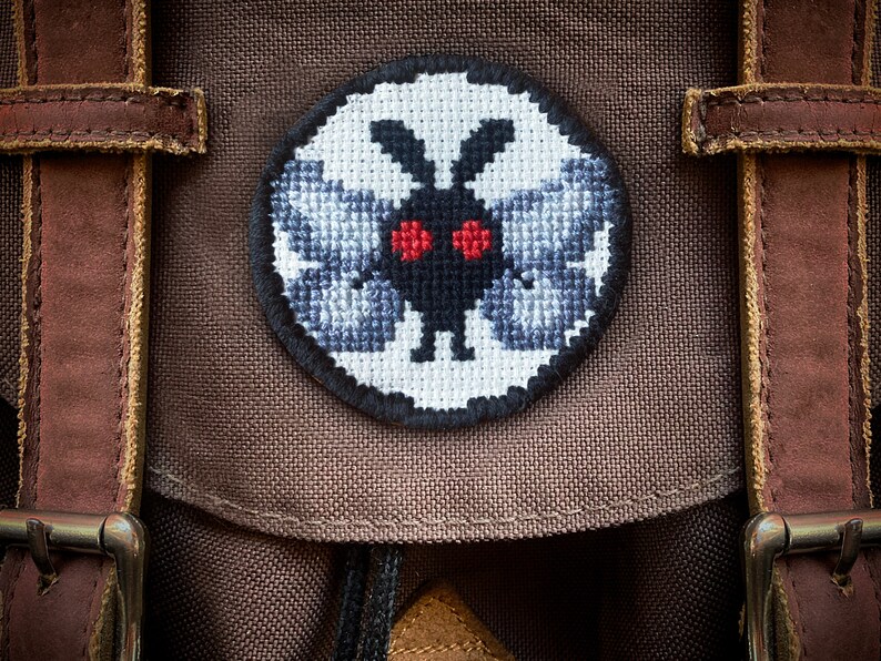 May include: A brown canvas backpack with a circular embroidered patch featuring a black and white moth with red eyes.