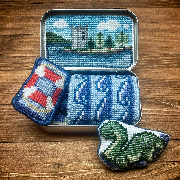 Puede incluir: Una lata de metal con una imagen bordada en azul y blanco de un castillo en un lago. La lata contiene tres pequeños cojines bordados: un salvavidas rojo y blanco, un cojín a rayas azul y blanco y un dinosaurio verde y blanco.