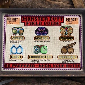 May include: A framed cross-stitch sampler titled Monster Butt Field Guide, featuring six illustrated monster posteriors including a zombie, Dracula, mummy, ghost, Frankenstein, and werewolf, with text instructions and locations on a light fabric.