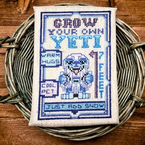 May include: Cross-stitch art piece featuring a blue and white yeti design with the text "Grow Your Own Yeti." The artwork includes the phrases "Warm Hugs," "Cool Pet," and "Just Add Snow." The piece is displayed in a woven basket.