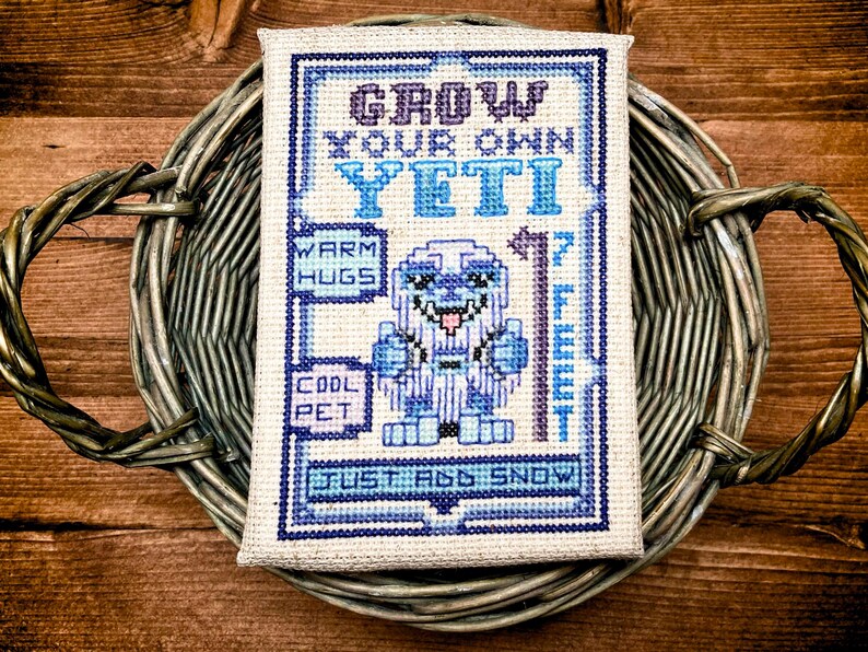 May include: Cross-stitch art piece featuring a blue and white yeti design with the text "Grow Your Own Yeti." The artwork includes the phrases "Warm Hugs," "Cool Pet," and "Just Add Snow." The piece is displayed in a woven basket.