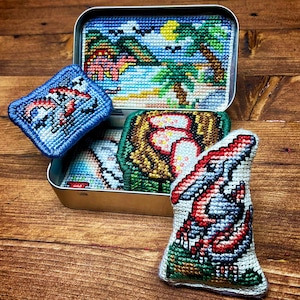 May include: A metal tin containing several cross-stitched items. The tin's lid features a tropical beach scene. Other items include a blue square with a dinosaur, and a larger dinosaur-shaped item. The items are on a wooden surface.