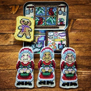 Może przedstawiać: Kolekcja ręcznie robionych ozdób świątecznych wykonanych ściegiem igłowym. Zestaw zawiera trzy figurki Pani Mikołajowej, piernikowego ludzika i ozdobną puszkę ze sceną kuchenną. Ozdoby są kolorowe i szczegółowe, idealne na święta.