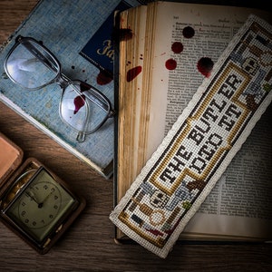 May include: A vintage book with a blood-stained bookmark that reads "The Butler Did It". The bookmark is a cross-stitch design with a brown and white color scheme. A pair of glasses and a small clock are also in the image.