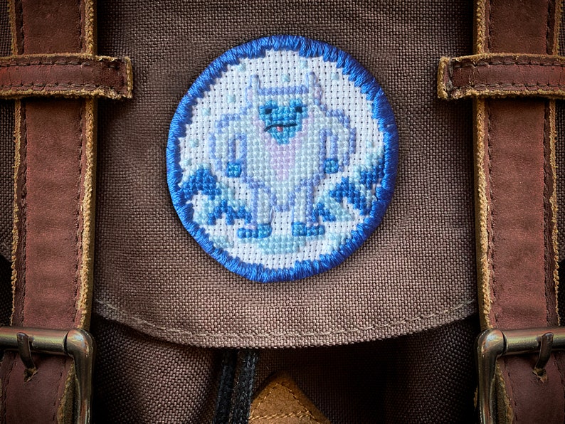 May include: A brown canvas backpack with a blue and white embroidered patch of a yeti in a snowy scene.