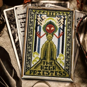 May include: A cross-stitch embroidery of the High Priestess tarot card. The card features a woman in a green robe with a crown and a red face. The background is blue and white.