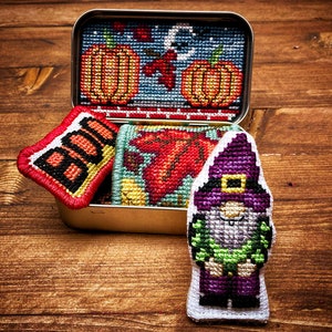 May include: A small tin box filled with four embroidered felt ornaments. The ornaments are a pumpkin, a leaf, a gnome, and a sign that says "BOO".