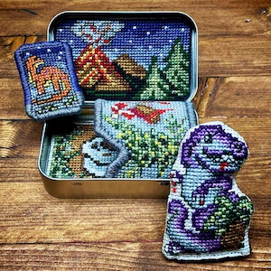 Może przedstawiać: Kolekcja ręcznie robionych przedmiotów haftowanych krzyżykiem, w tym małe metalowe pudełko z krajobrazem, prostokątna naszywka z dinozaurem i pluszowy dinozaur. Pudełko przedstawia wulkan i drzewa. Pluszowy dinozaur jest fioletowy i zielony.