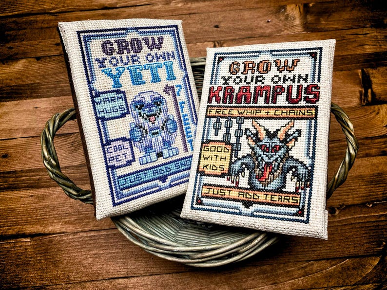 May include: Two cross-stitch samplers in a woven tray. One says "Grow Your Own Yeti" with a cartoon yeti. The other says "Grow Your Own Krampus" with a Krampus illustration. Both have text and are framed in blue.