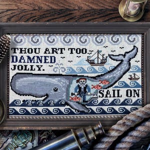 Puede incluir: Un diseño de punto de cruz enmarcado que presenta una ballena con un marinero en su espalda. La ballena es azul y gris, y el marinero lleva un sombrero rojo y una camisa azul. El texto "Thou art too damned jolly. Sail on" está cosido en hilo negro.