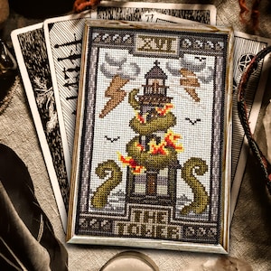 May include: A cross-stitch embroidery of the Tower card from a tarot deck. The card depicts a tower with flames and a snake wrapped around it. The card is framed in a wooden frame.