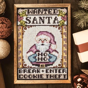 May include: A framed cross-stitch featuring Santa Claus holding a sign that says "HO HO" and the text "WANTED SANTA" and "BREAK + ENTER COOKIE THEFT". The image is surrounded by Christmas ornaments and a wrapped gift.