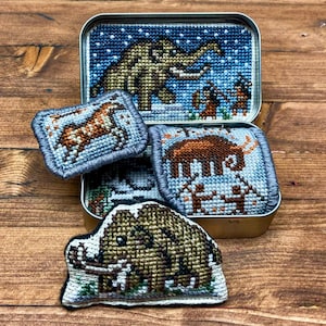 May include: A selection of cross-stitched patches and a tin, featuring prehistoric animal designs. The tin lid shows a woolly mammoth and figures, with patches depicting a deer, a mammoth, and another mammoth. The color palette includes brown, blue, and white.