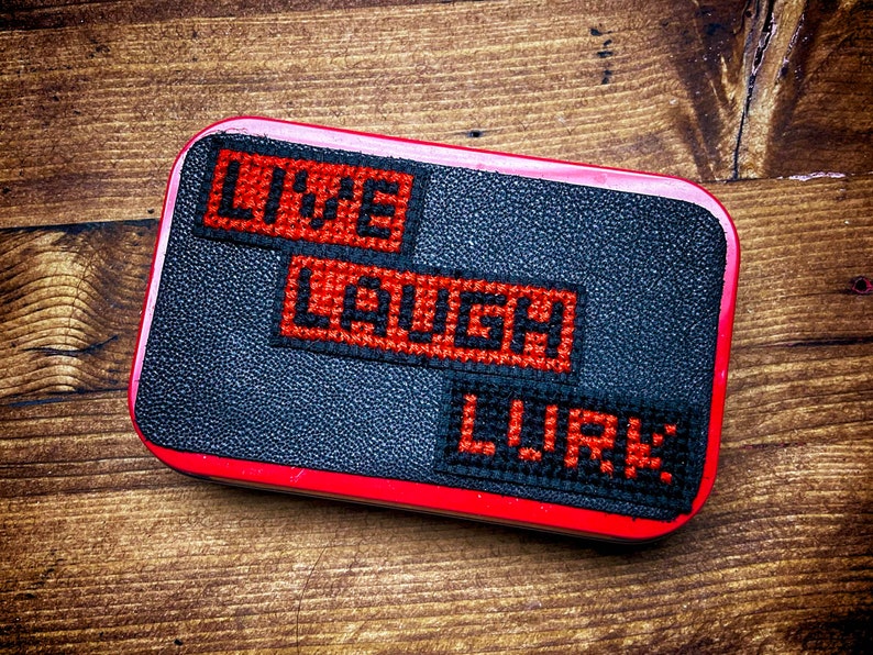 Op de afbeelding: Een zwarte blik met een rode rand en drie rode en zwarte geborduurde pleisters die "LIVE", "LAUGH" en "LURK" zeggen.