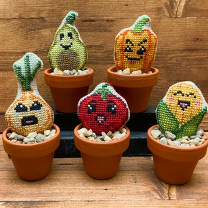 May include: Five small potted plants with embroidered faces. The plants are an avocado, a pumpkin, an onion, a tomato, and corn. The plants are all in terracotta pots with small rocks at the bottom.
