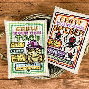 Könnte beinhalten: Zwei rechteckige Kreuzstichbilder auf hellem Stoff. Das linke zeigt eine Kröte mit violettem Hut und den Text GROW YOUR OWN TOAD, WITCHY PET, RIBET, CHANCE AT A PRINCE, JUST ADD EYE OF NEWT. Das rechte zeigt eine Spinne mit dem Text GROW YOUR OWN SPIDER, CREEPY AND CRAWLY, WEB DEVELOPER, JUST ADD FLIES.