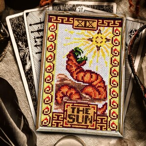 May include: A cross-stitch embroidery of The Sun tarot card. The card features a yellow sun with rays, a red and orange figure with a green crown, and the text "THE SUN" in a yellow banner.