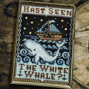 Puede incluir: Obra de arte de punto de cruz en un marco dorado con una ballena blanca, un barco y el texto "HAST SEEN THE WHITE WHALE?". El fondo es negro con estrellas amarillas. La obra está sobre un mapa antiguo.