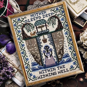 Puede incluir: Arte de punto de cruz enmarcado. La obra presenta una escena de pozo con el texto "MOTHER SLEEPS DEEP WITHIN THE WISHING WELL". El diseño incluye una figura de esqueleto, un pozo y un sauce llorón, con detalles en azul y verde.