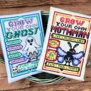 May include: Two cross-stitch samplers with humorous phrases. One says "Grow Your Own Ghost" with a ghost illustration. The other says "Grow Your Own Mothman" with a moth illustration. Both are in a woven basket.