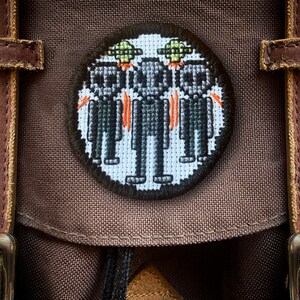 May include: A brown canvas backpack with a round embroidered patch featuring three pixelated figures wearing green hats and a flying saucer above them.