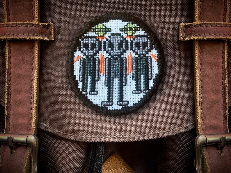 May include: A brown canvas backpack with a round embroidered patch featuring three pixelated figures wearing green hats and a flying saucer above them.